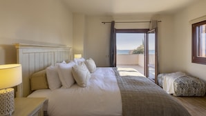 Een slaapkamer met een groot bed, een nachtkastje, een lamp, uitzicht op zee door een geopende deur en een raam met gordijnen.