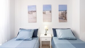 Ein Hotelzimmer mit zwei Betten, einem Nachttisch mit Lampe und drei Wandbildern zum Thema Strand.
