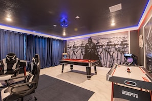 A room with a pool table, two gaming chairs, a foosball table, and a Star Wars-themed wall.