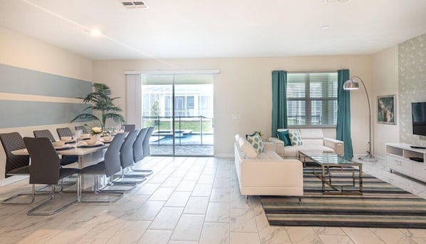A modern living room with a dining area, a sofa set, a TV, and a balcony with a pool view.
