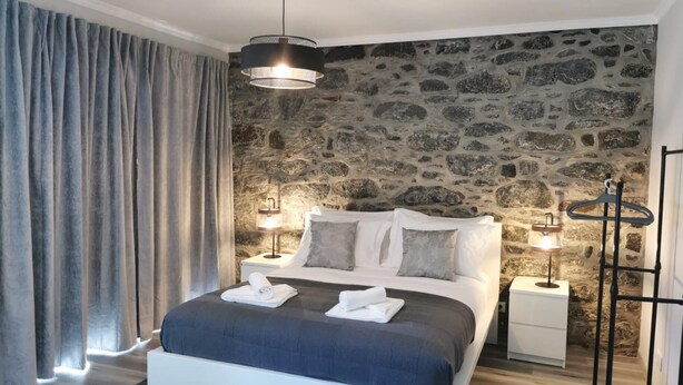 Un dormitorio moderno con pared de piedra, una cama grande con ropa de cama blanca, dos mesitas de noche con lámparas y un perchero negro.