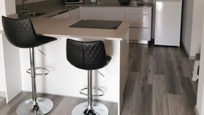 Una cocina moderna con una isla, dos taburetes de bar, una campana extractora de acero inoxidable, microondas, hervidor, fregadero, refrigerador y un cuadro en la pared.