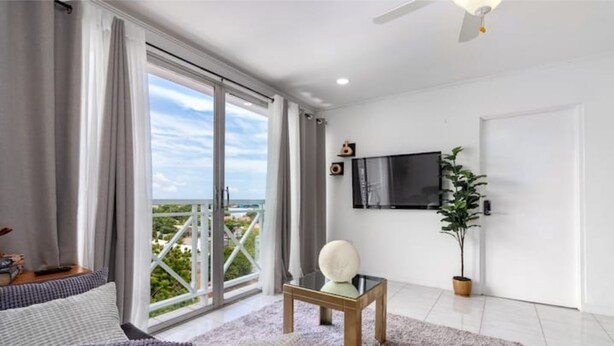 Een moderne woonkamer met een grote schuifpui naar een balkon, een flatscreen televisie aan de muur, een tafeltje met een decoratief object en een potplant.