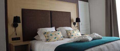 A neatly made bed with white bedding and teal throw pillows, a wooden nightstand with a lamp, and a wall-mounted light fixture.