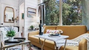 Ein gemütliches Wohnzimmer mit einer Couch, einem kleinen Tisch, einer Topfpflanze und einem Fenster mit Blick auf Bäume.