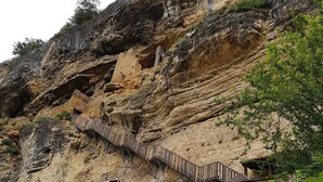 Ein Steinhaus mit einer Holztreppe, die einen steilen Klippenhang hinaufführt, umgeben von Grün.