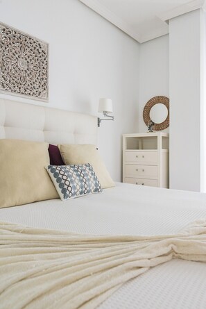 Une chambre à coucher bien rangée, avec un lit, des oreillers, une lampe fixée au mur, un miroir décoratif et un meuble de rangement.