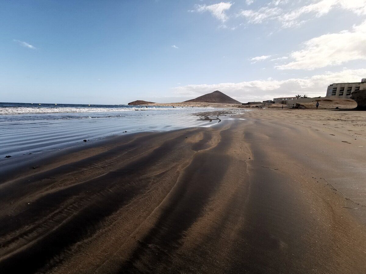 Ein Strand mit einem Berg im Hintergrund, sandigem Gelände und blauem Himmel.