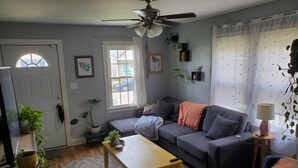 A living room with a sofa, a coffee table, and a ceiling fan. There are windows with curtains, a door, and a plant on a stand. - Short Walk to Churchill Downs! (Louisville)