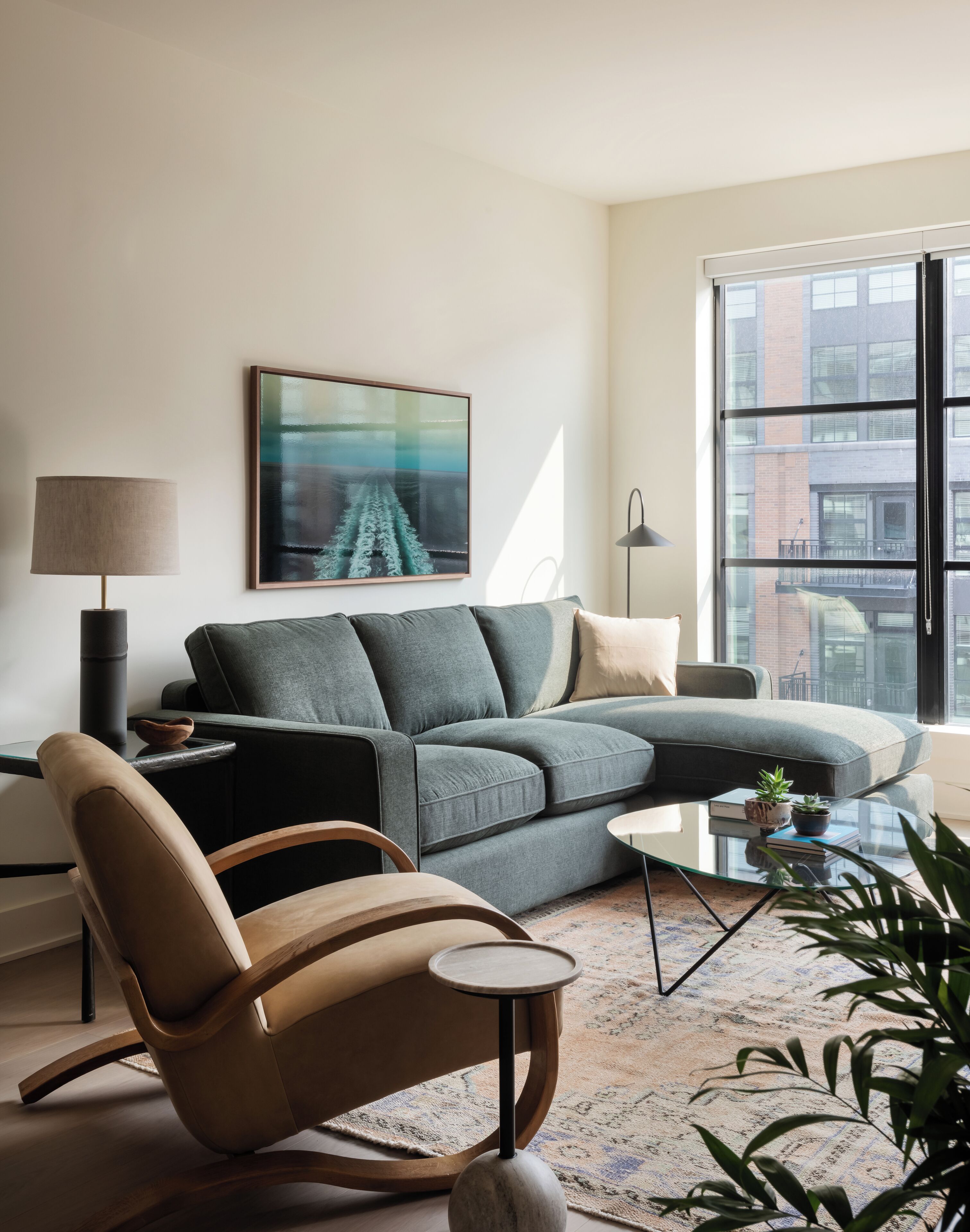 A modern living room with a sectional sofa, a glass coffee table, a wooden chair, a floor lamp, and a large window with a city view.