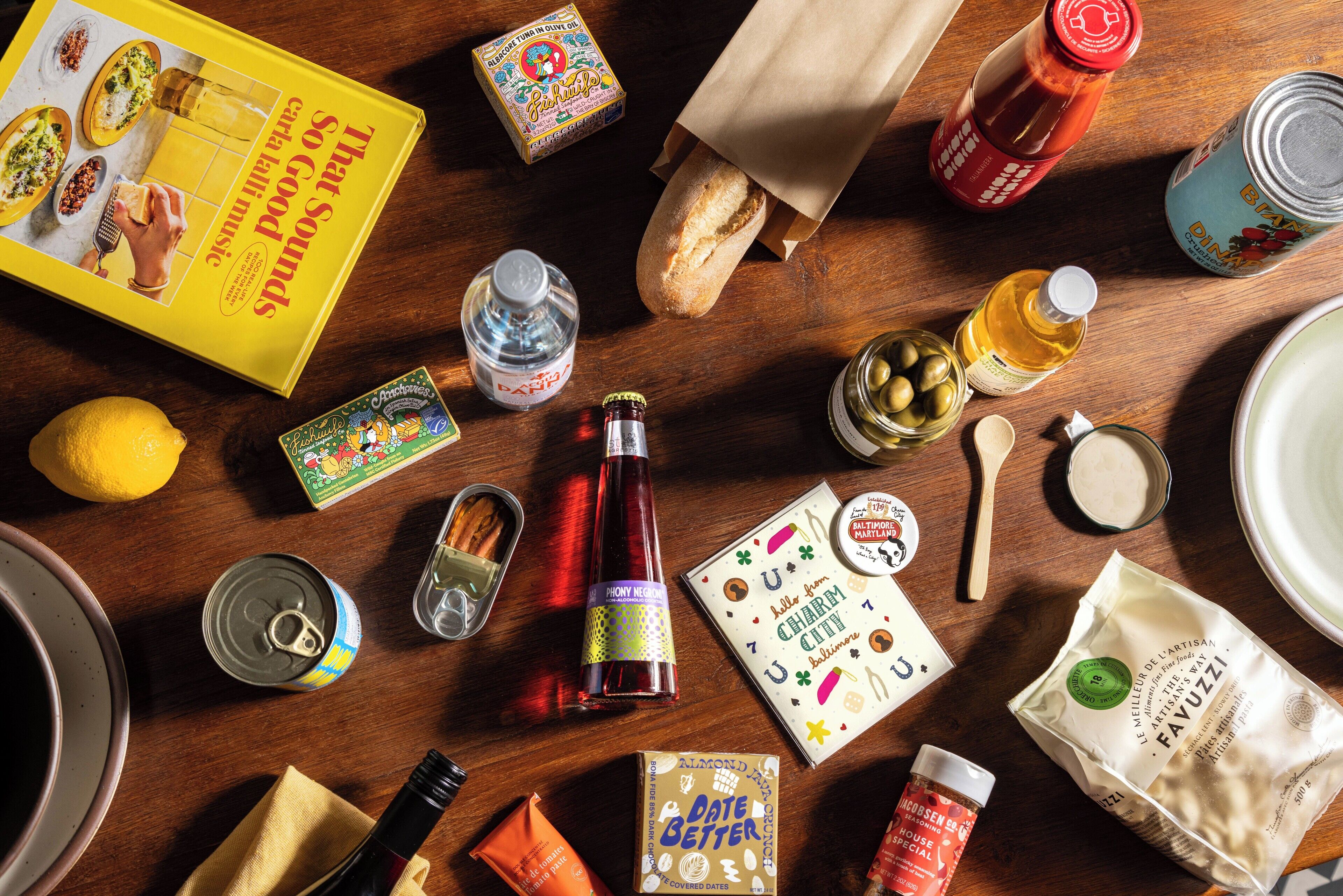 A variety of food items and condiments on a wooden table, including a book, a bottle of sauce, a lemon, and a can of soda.