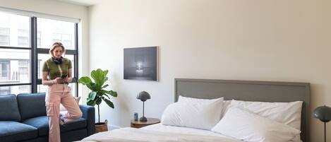 A modern bedroom with a bed, a sofa, a large window, and a plant.