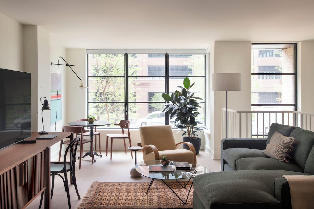 A modern living room with a sofa, armchair, and a dining table with chairs. There is a TV on a wooden cabinet, a floor lamp, and a large window with a view of buildings and trees.
