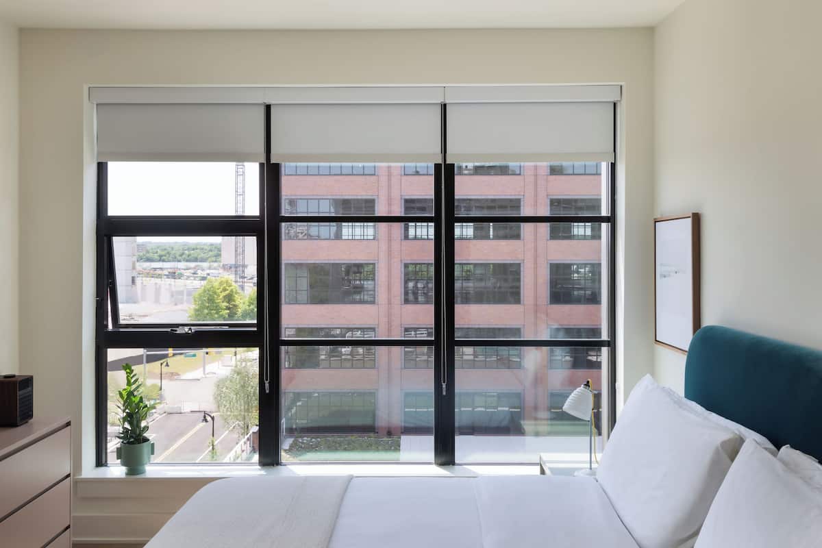 A bedroom with a large window, a bed, a bedside table, a lamp, and a framed picture on the wall.