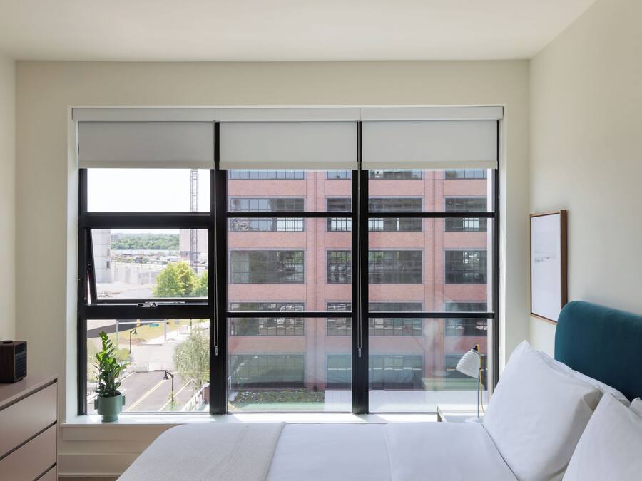 A bedroom with a large window, a bed, a bedside table, a lamp, and a framed picture on the wall.