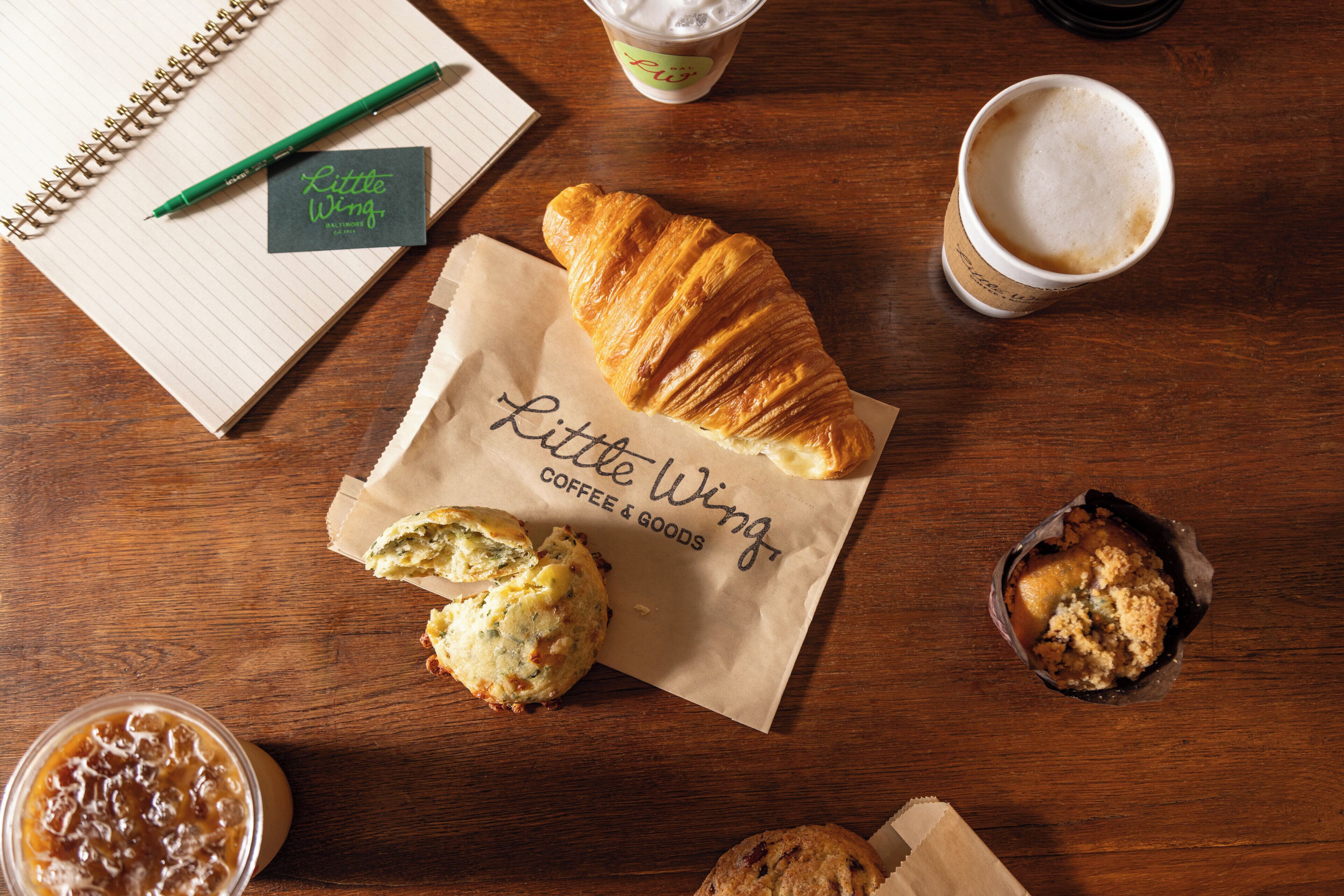 A wooden table with a notebook, a pen, a croissant, a muffin, and a cup of coffee.