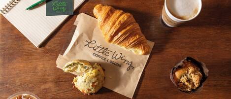 Una mesa de madera con un cuaderno, un bolígrafo, un croissant, un muffin y una taza de café.