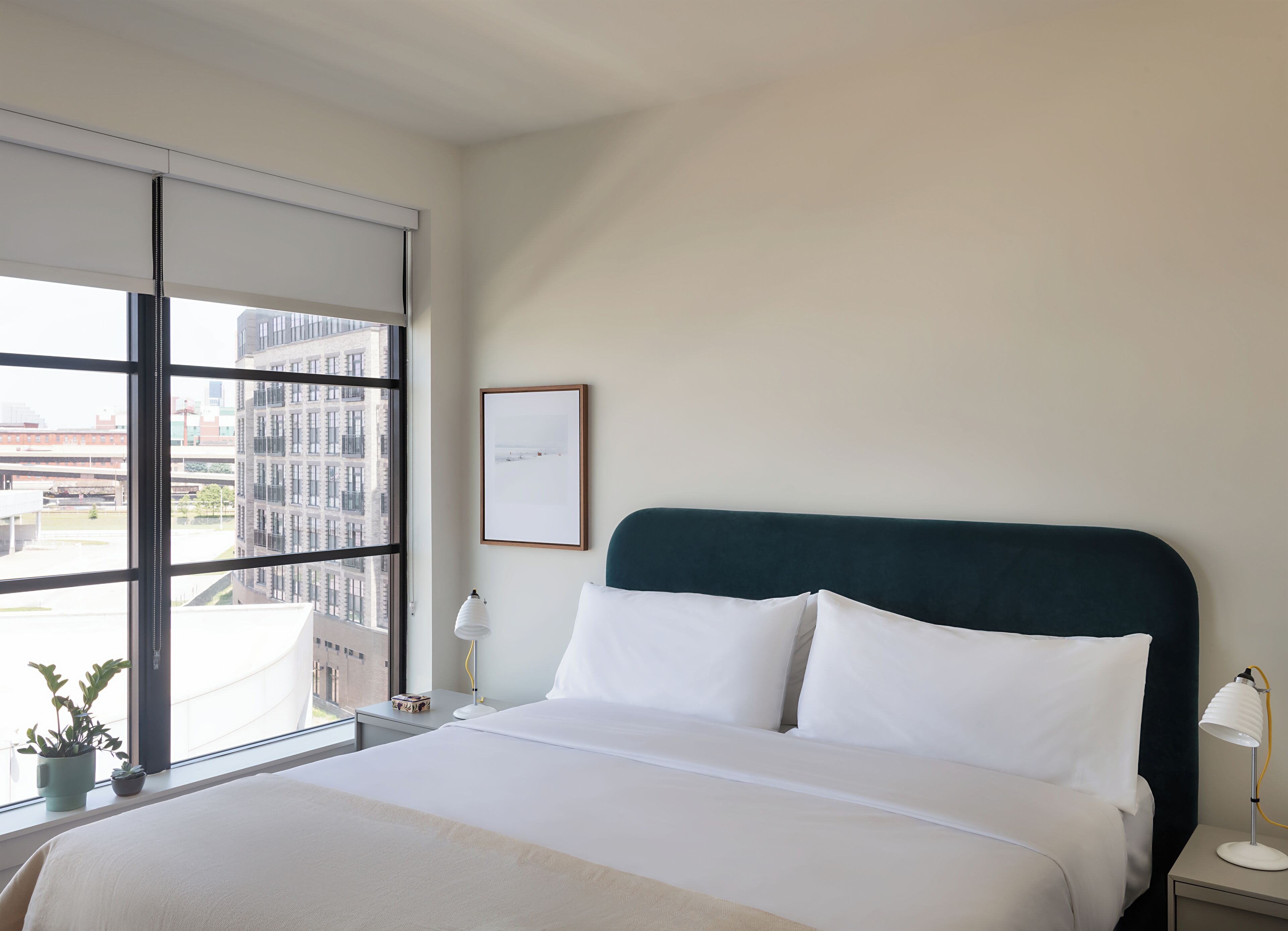 Une chambre d’hôtel moderne avec un grand lit, une vue sur la ville grâce à de grandes fenêtres du sol au plafond, un tableau encadré au mur et une petite table avec une lampe.