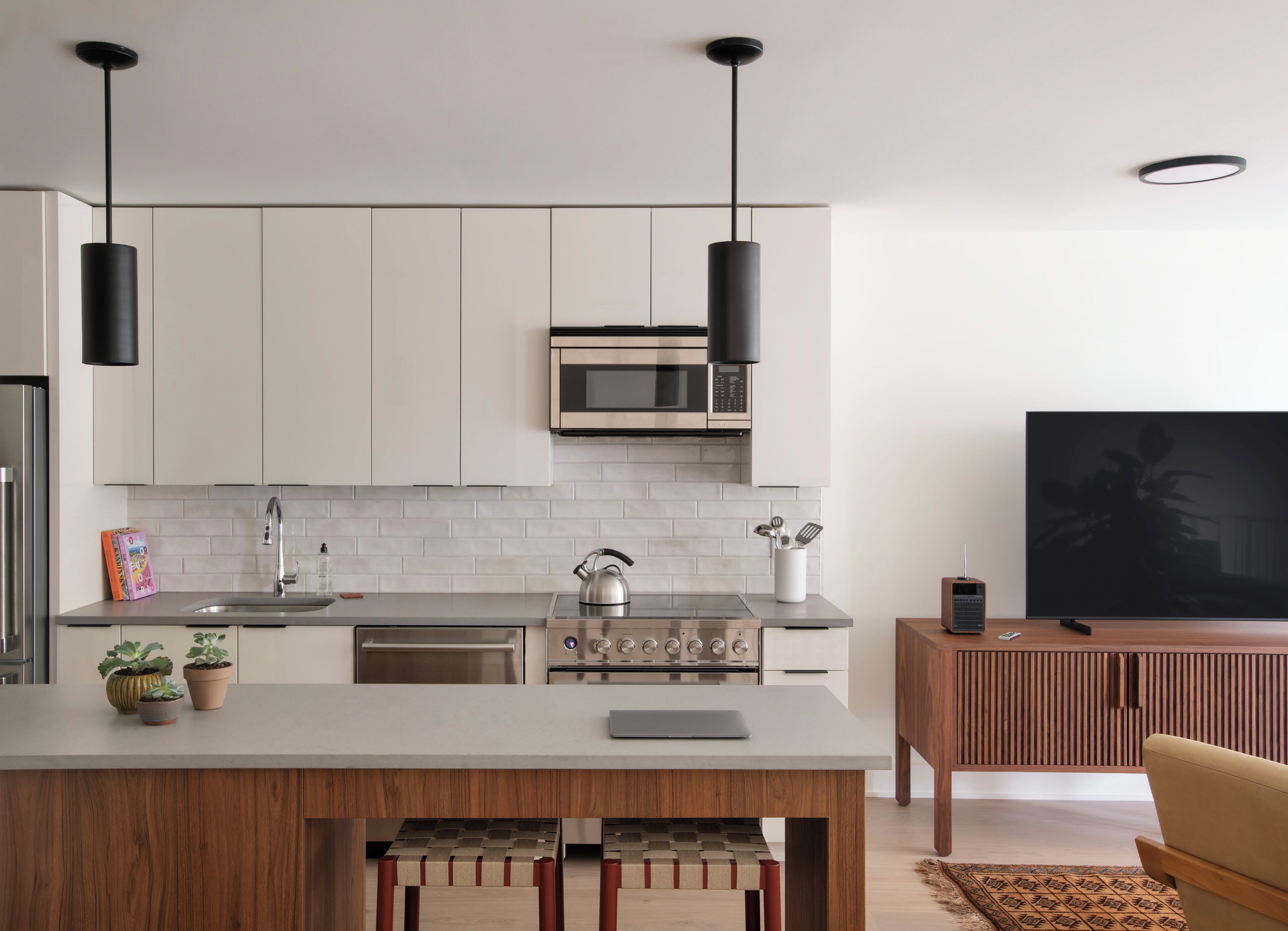 A modern kitchen with white cabinets, a stainless steel stove, and a microwave. There is a wooden island with a kettle and a tablet. A TV is mounted on the wall, and a plant is visible in the corner.