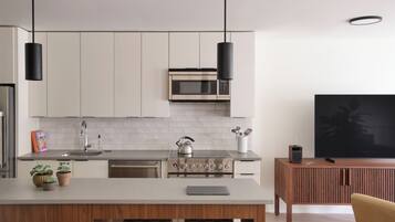 A modern kitchen with white cabinets, a stainless steel stove, and a microwave. There is a wooden island with a kettle and a tablet. A TV is mounted on the wall, and a plant is visible in the corner.