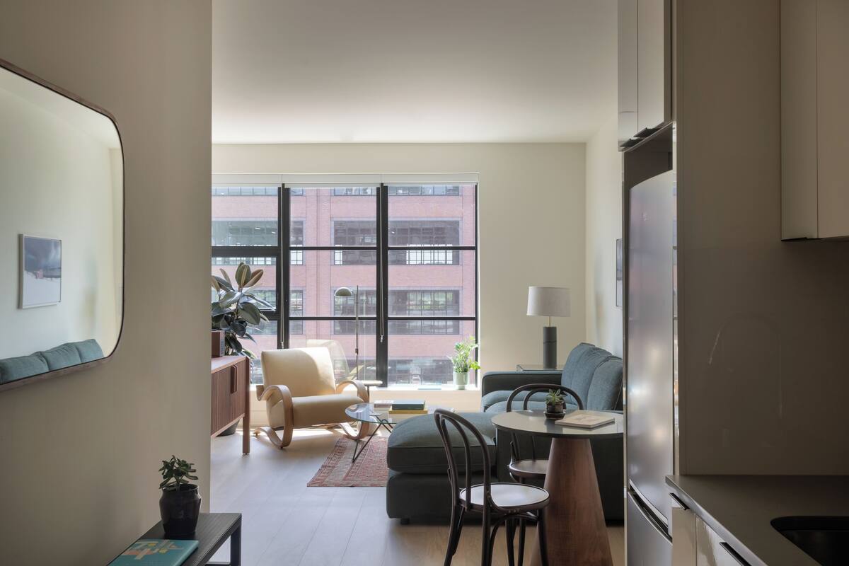 A modern living room with a sofa, a dining table with chairs, a small side table with a lamp, and a large window with a view of a brick building.