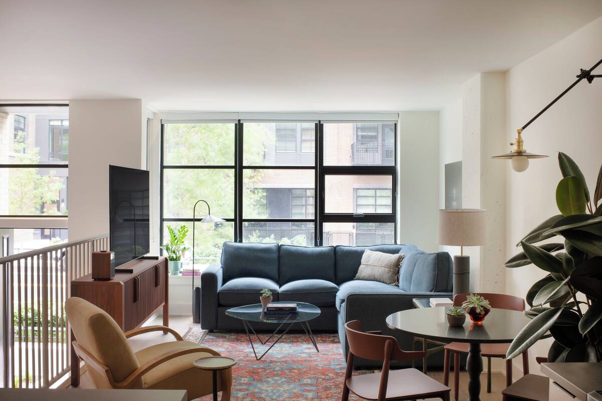 A modern living room with a sofa, coffee table, chairs, and a TV. There are large windows with a view of buildings and trees.
