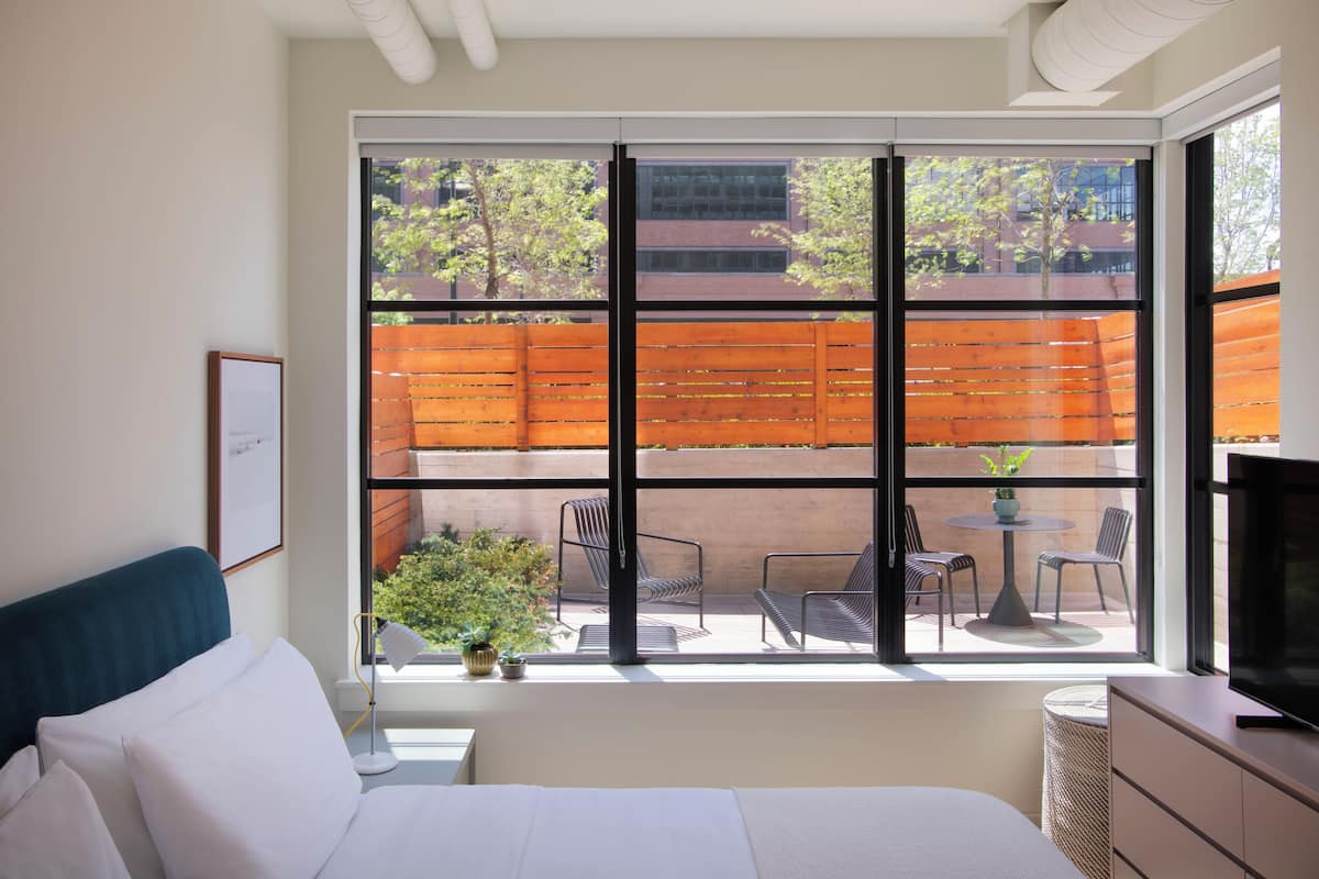 A bedroom with a large window, a bed, a bedside table, a lamp, and a TV.