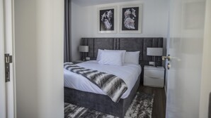 Ein modernes Hotelzimmer mit einem großen Bett, zwei Nachttischen, einem grauen Kopfteil und zwei gerahmten floralen Kunstwerken an der Wand.