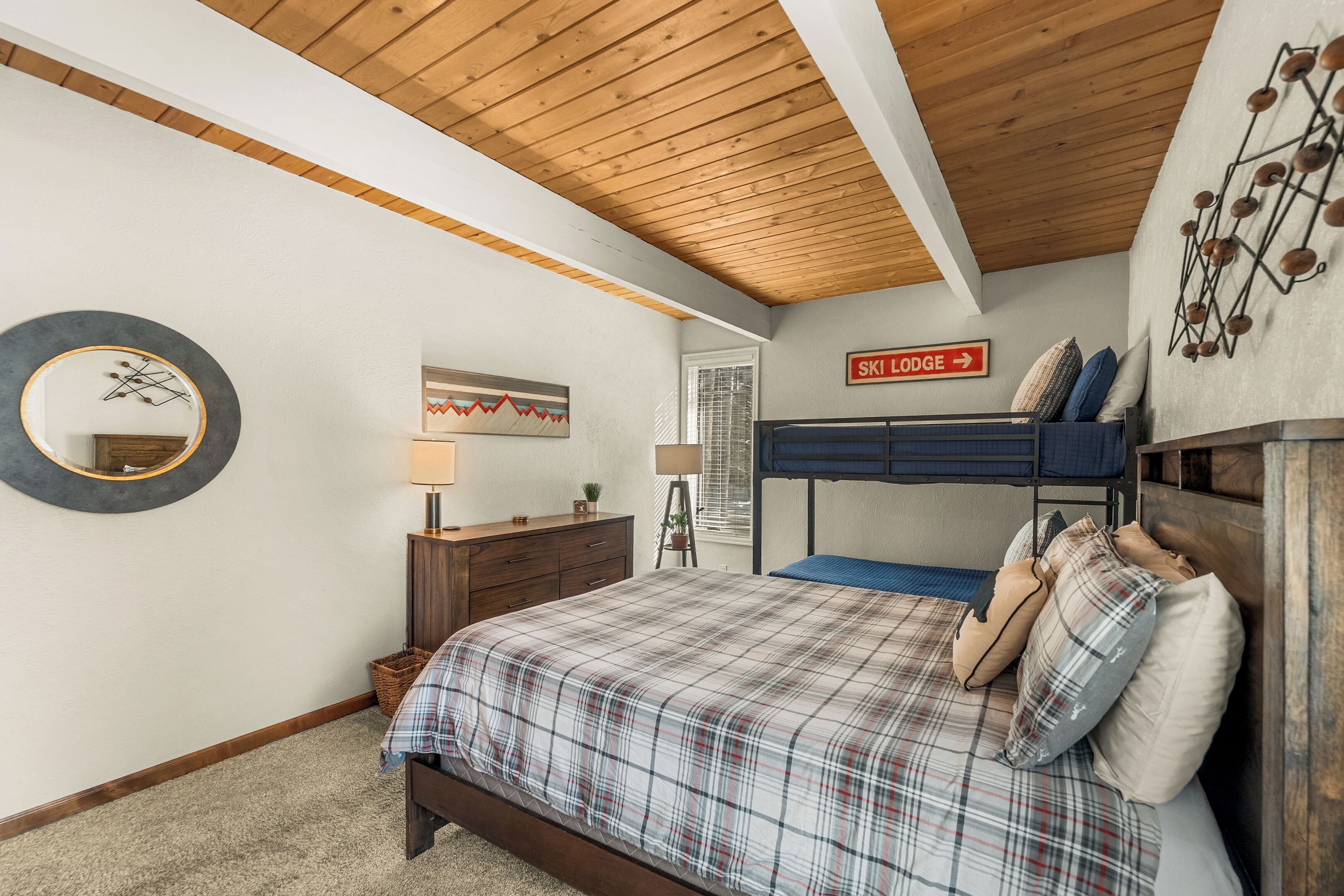 A bedroom with a wooden ceiling, a bunk bed, a plaid bedspread, a mirror, a dresser, and a sign indicating a ski lodge direction.