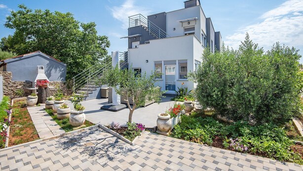 En moderne bygning i to etager med et overdækket udendørs område, stenplantere og forskellige planter.