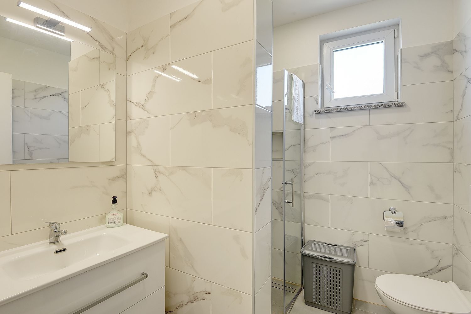 A bathroom with a marble tile backsplash, a white sink with a mirror, a glass shower enclosure, and a toilet.