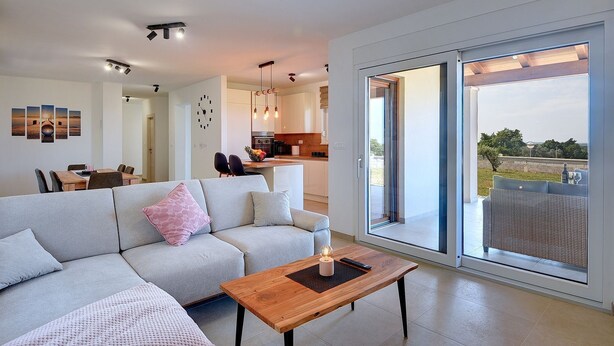 En moderne stue med sofa, sofabord og spiseplads med stole og bord. Et køkken er synligt i baggrunden, og en stor skydedør fører ud til et udendørs område.