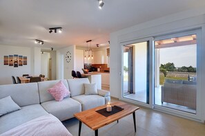 Uma sala de estar moderna com sofá, mesa de centro e área de jantar com cadeiras e mesa. Uma cozinha é visível ao fundo, e uma grande porta de vidro deslizante dá acesso a uma área externa.
