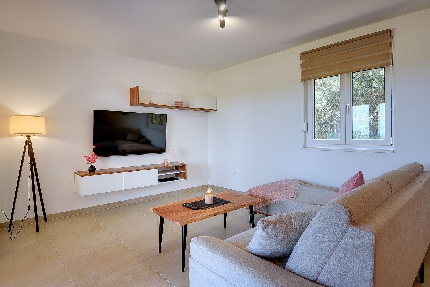 Una sala de estar moderna con un televisor de pantalla plana montado en una pared blanca, una mesa de centro de madera, un sofá gris con cojines, una lámpara de pie y una ventana con persianas.