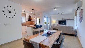 Et moderne køkken med en træø, et spiseområde med et langt bord og stole, en opholdsstue med sofa og fjernsyn, samt et stort vægur.