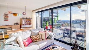A modern living room with a large window, a sofa with multiple cushions, a dining area with a wooden table, and a decorative Buddha statue. - Exclusive, quiet urbanization Oasis 325 (AN)