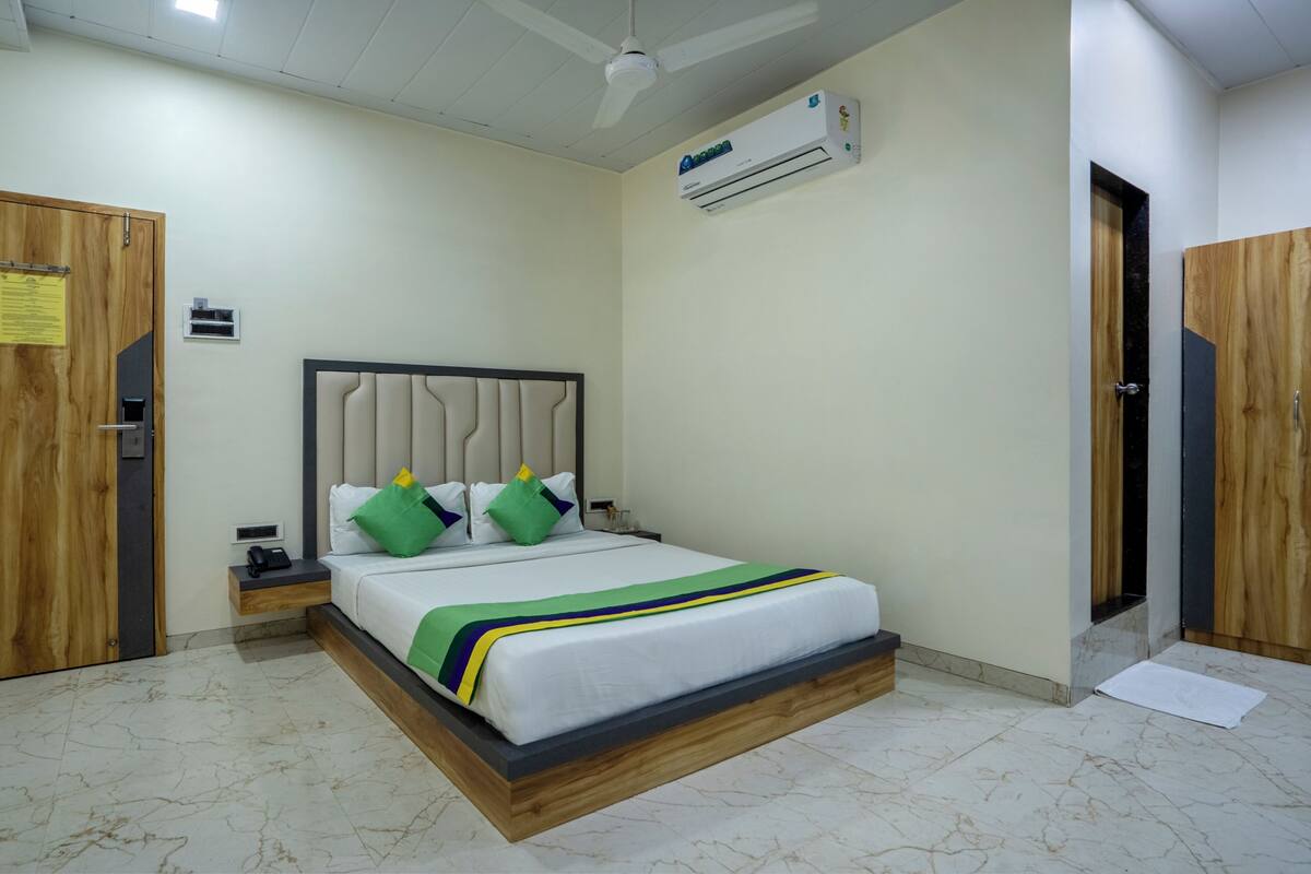 A hotel room with a bed, wooden wardrobe, an air conditioning unit, a ceiling fan, and a small bedside table with a phone.