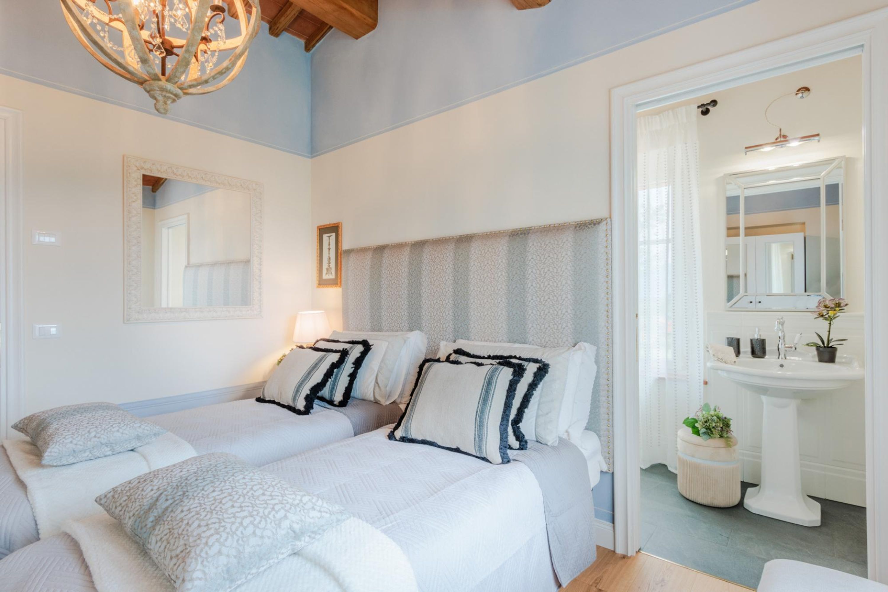 A bedroom with two beds, a large mirror, a chandelier, and a bathroom with a sink and mirror.