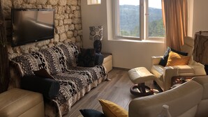 A living room with a stone wall, a flat-screen TV, a patterned sofa, a chair, and a view of the outdoors. - Maison de Village Située Dans un Hameau Très Calme , Agréable à Vivre (saint julien du serre)