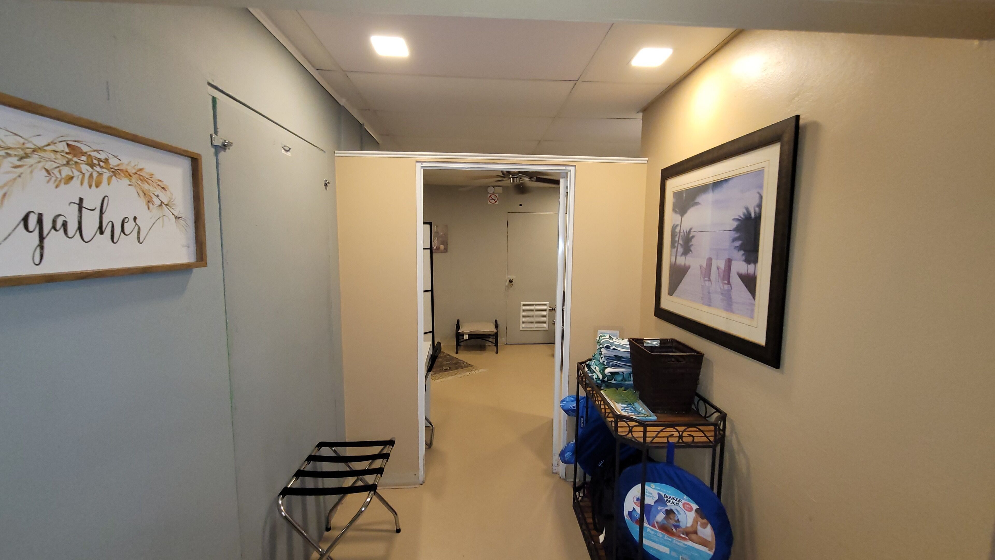 A hallway with a framed 'gather' sign, a chair, a small table, and a rack with folded towels.
