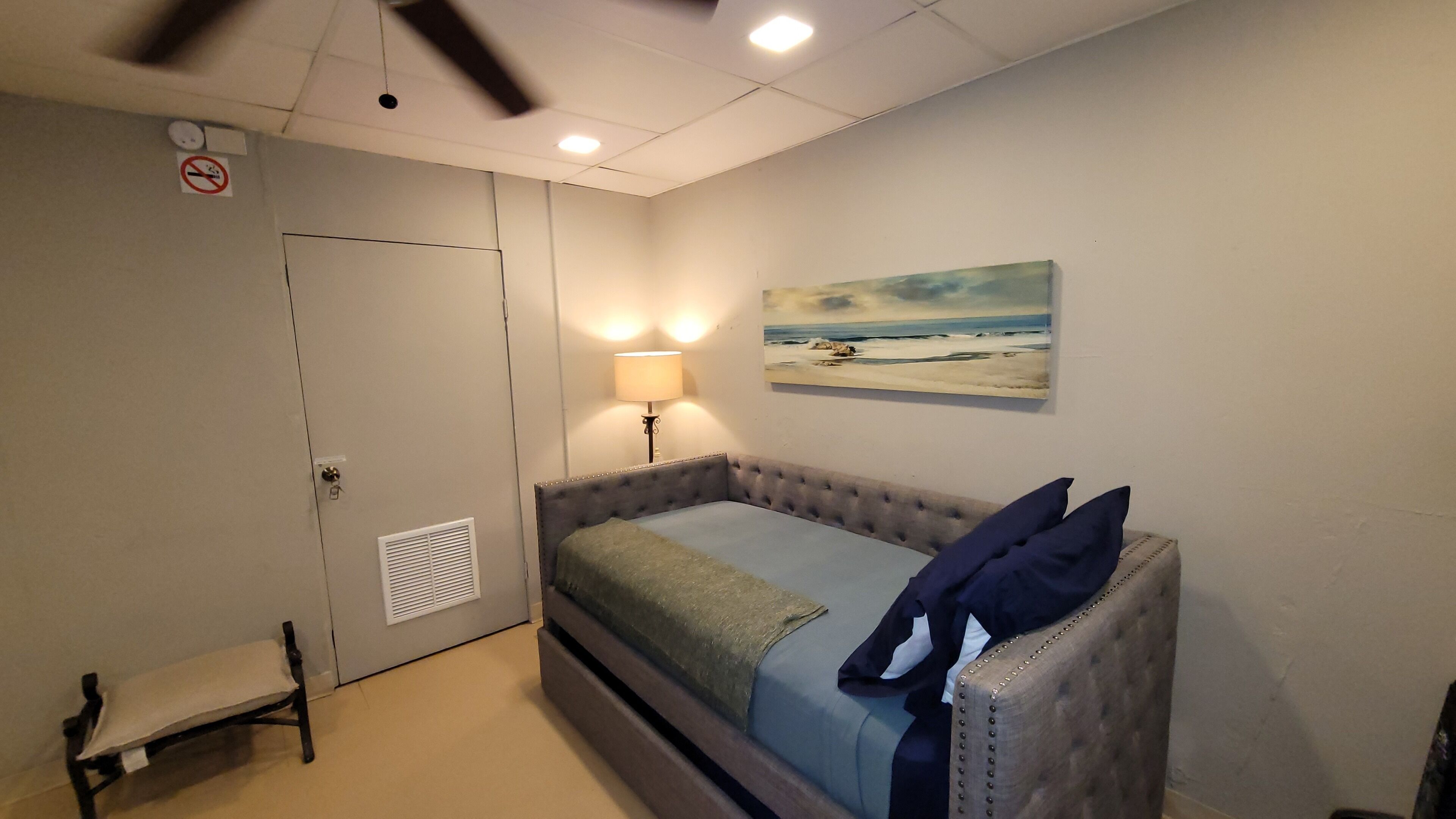 A bedroom with a bed, a bench, a wall-mounted heater, a ceiling fan, a painting, and a door with a no-smoking sign.