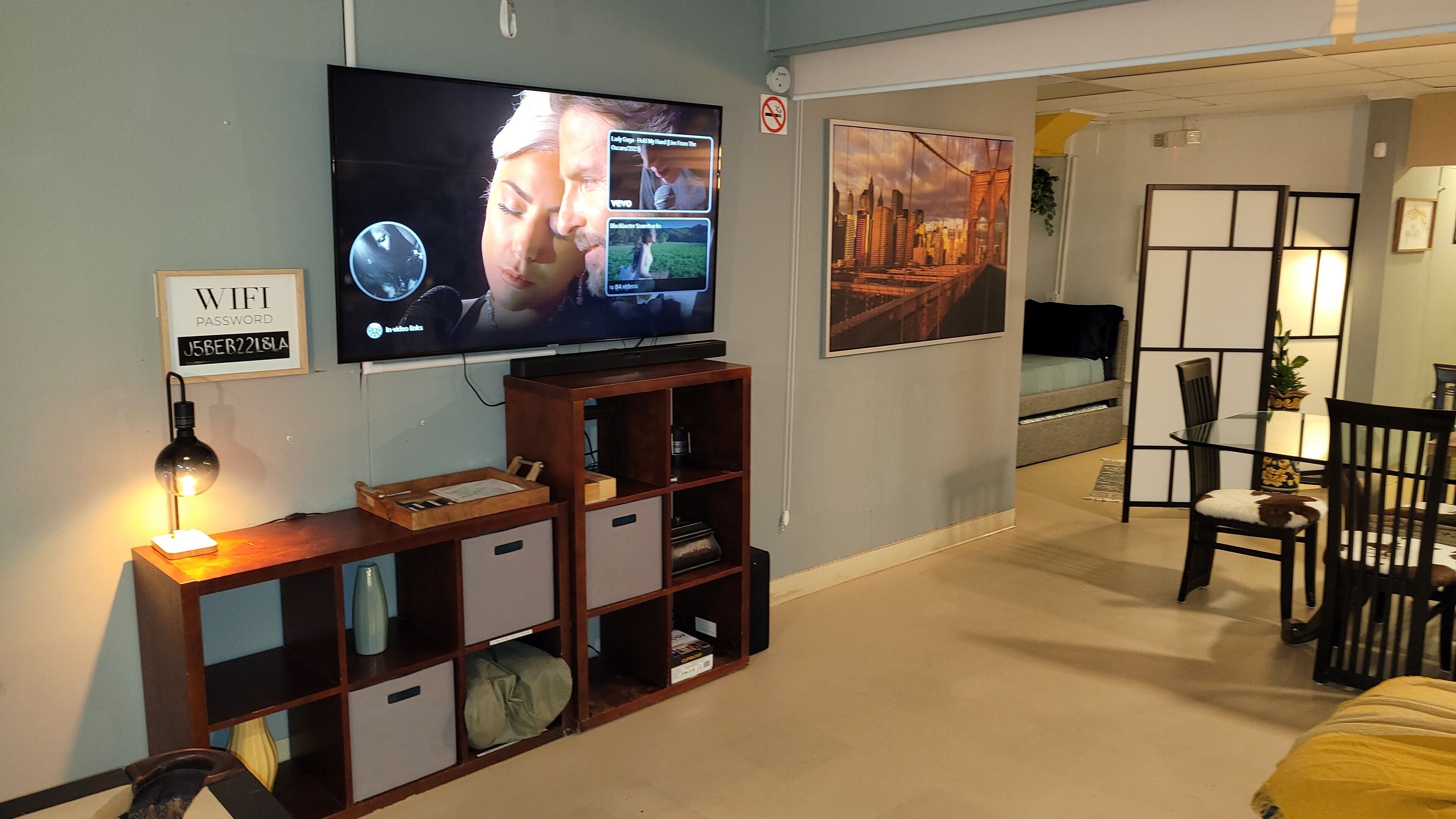 Una sala de estar con televisor, un mueble bajo para equipo audiovisual, una zona de comedor con mesa y sillas, y una pared decorada con marcos de fotos.