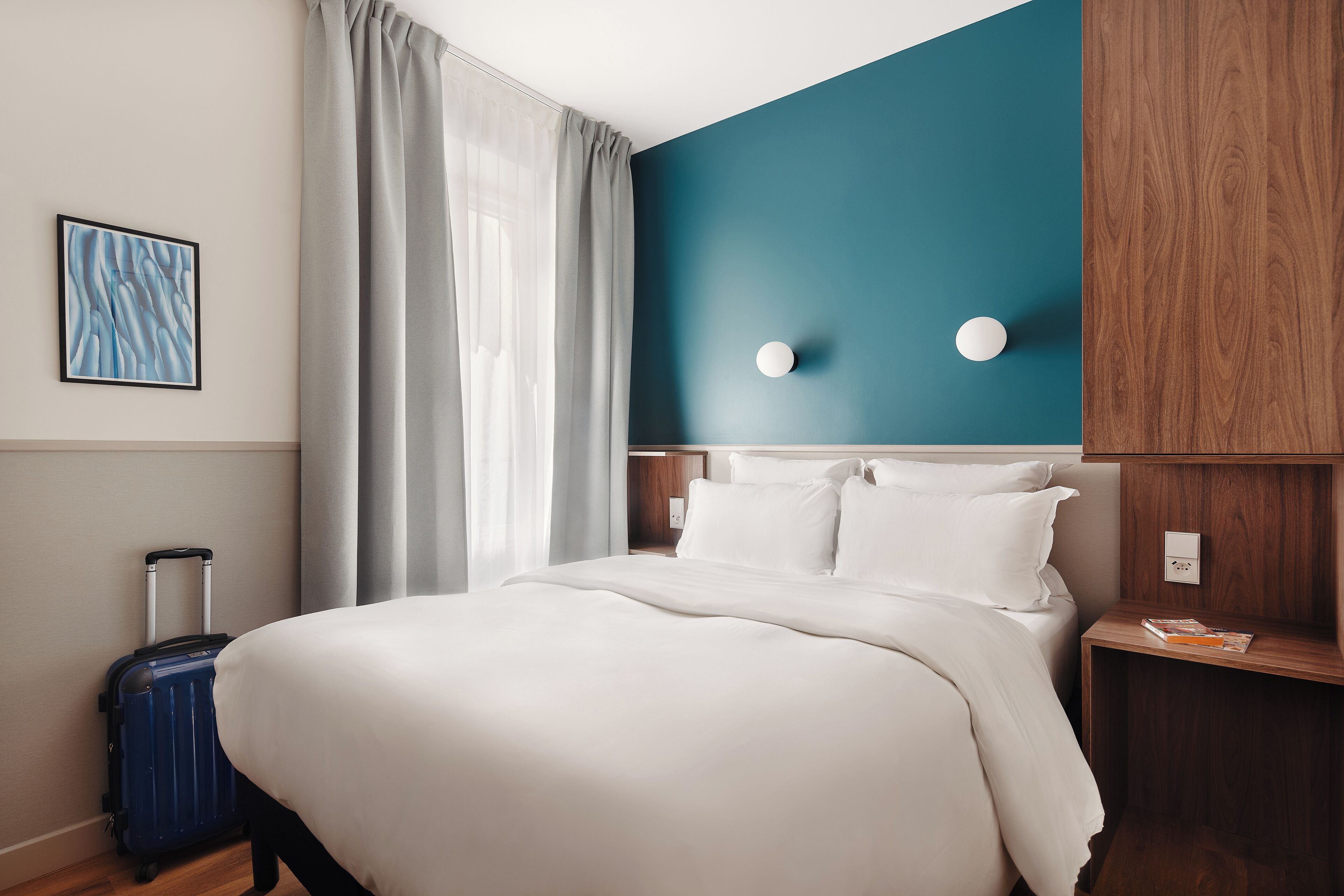 A hotel room with a bed, a suitcase, a painting on the wall, and a blue accent wall.