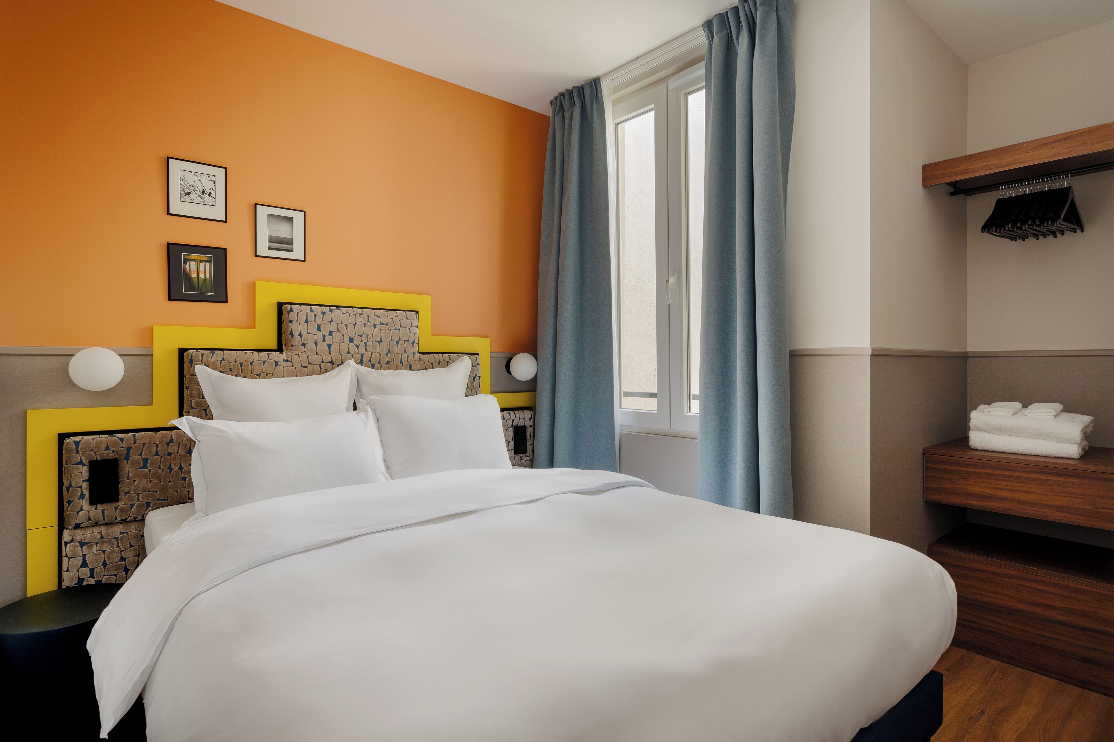 A hotel room with a bed, a wooden shelf with towels, a window with curtains, and wall-mounted artwork.