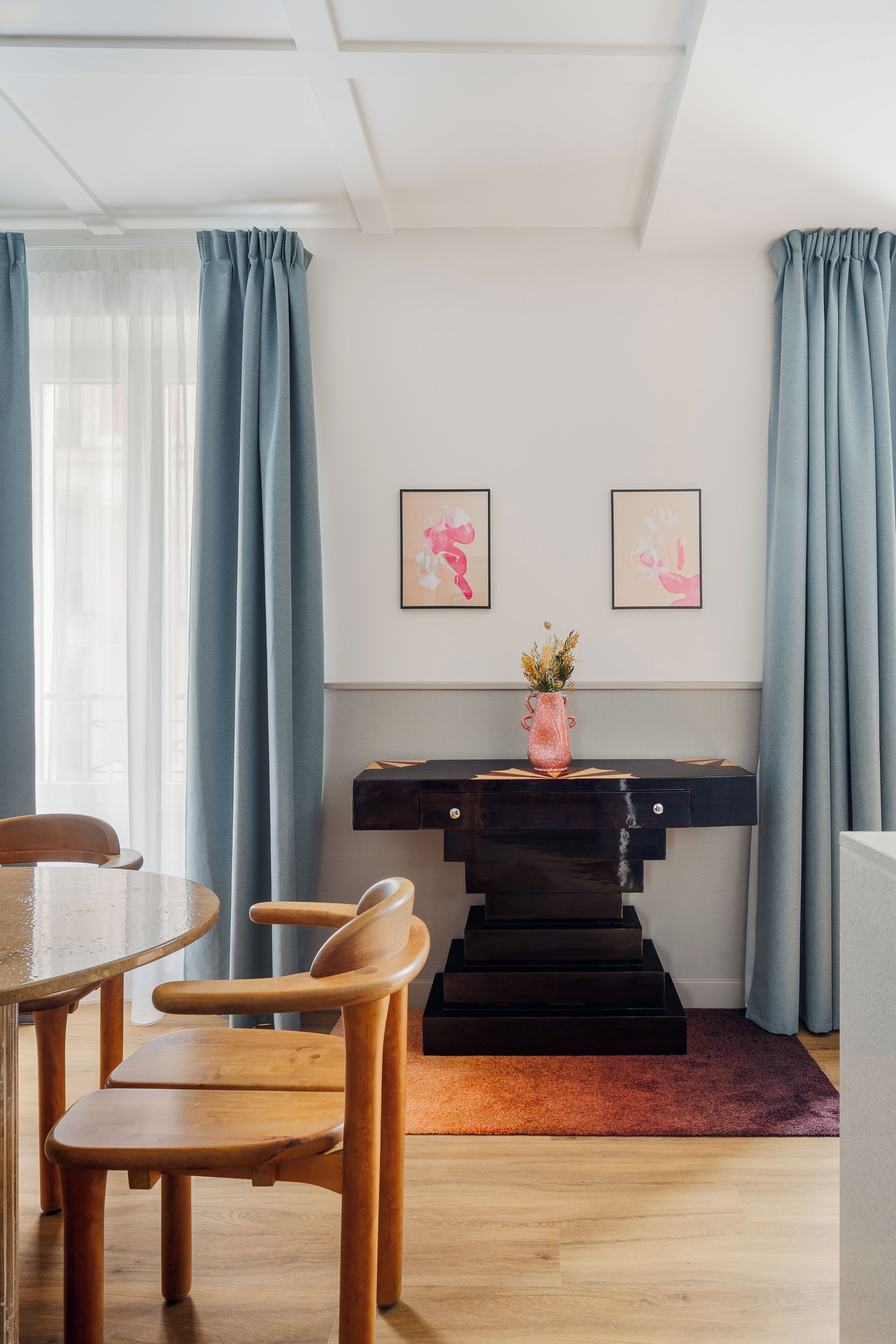 A modern room with a wooden dining table, chairs, a black console table with a vase, and artwork on the wall.