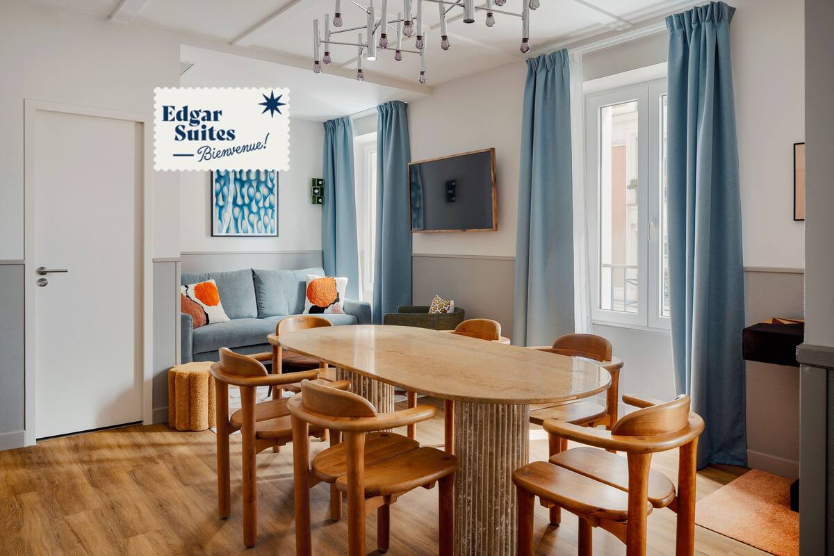 Een moderne eetruimte met een houten tafel en stoelen, een blauwe bank en een groot raam met gordijnen.
