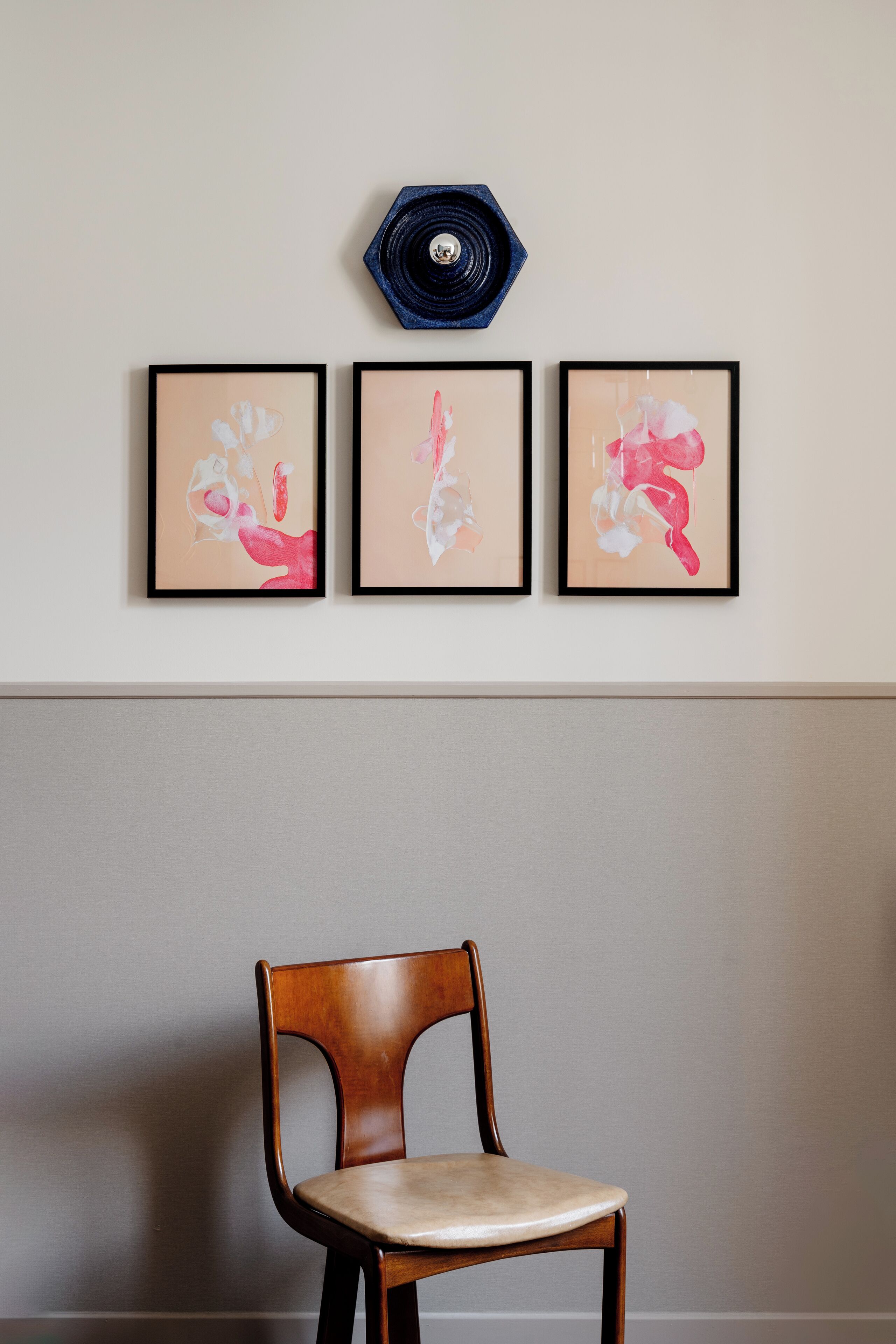 A wall with three framed abstract paintings and a wooden chair.