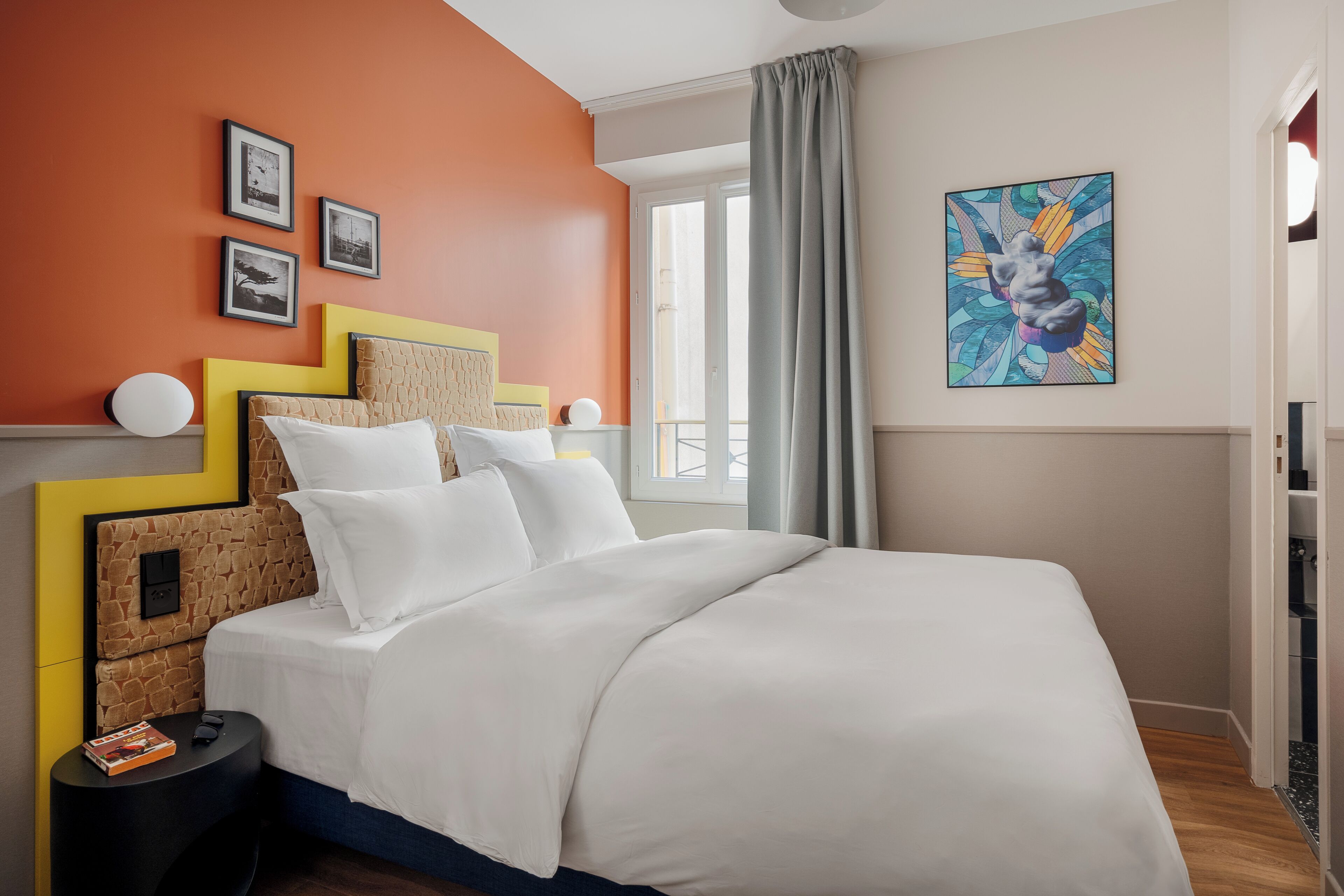 A modern bedroom with a bed, a nightstand, a window with curtains, and a wall with framed pictures.