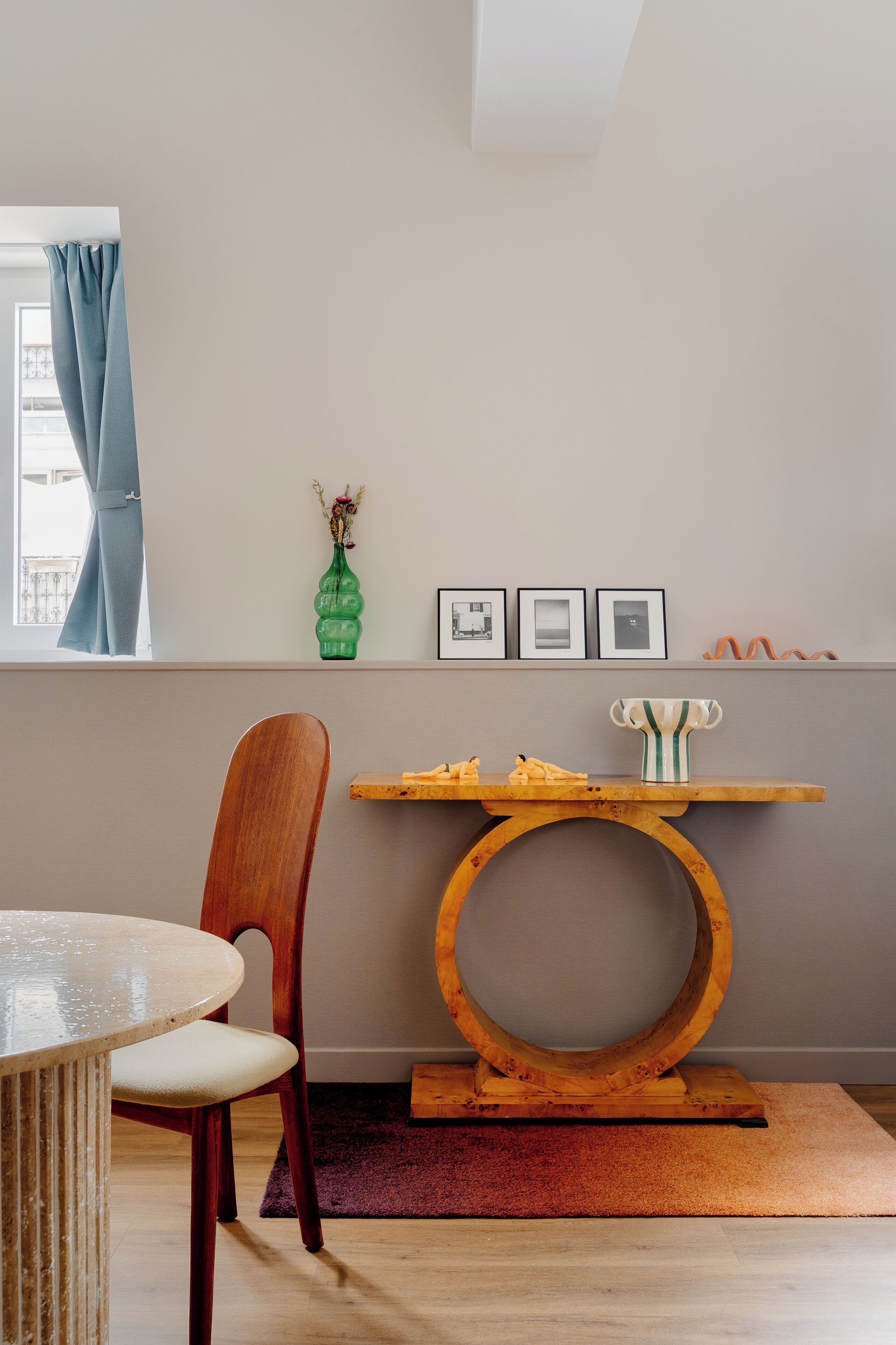 Une pièce comprenant une table à manger, une chaise en bois, une petite table ronde en bois, une étagère avec des objets décoratifs et une fenêtre ornée de rideaux bleus.