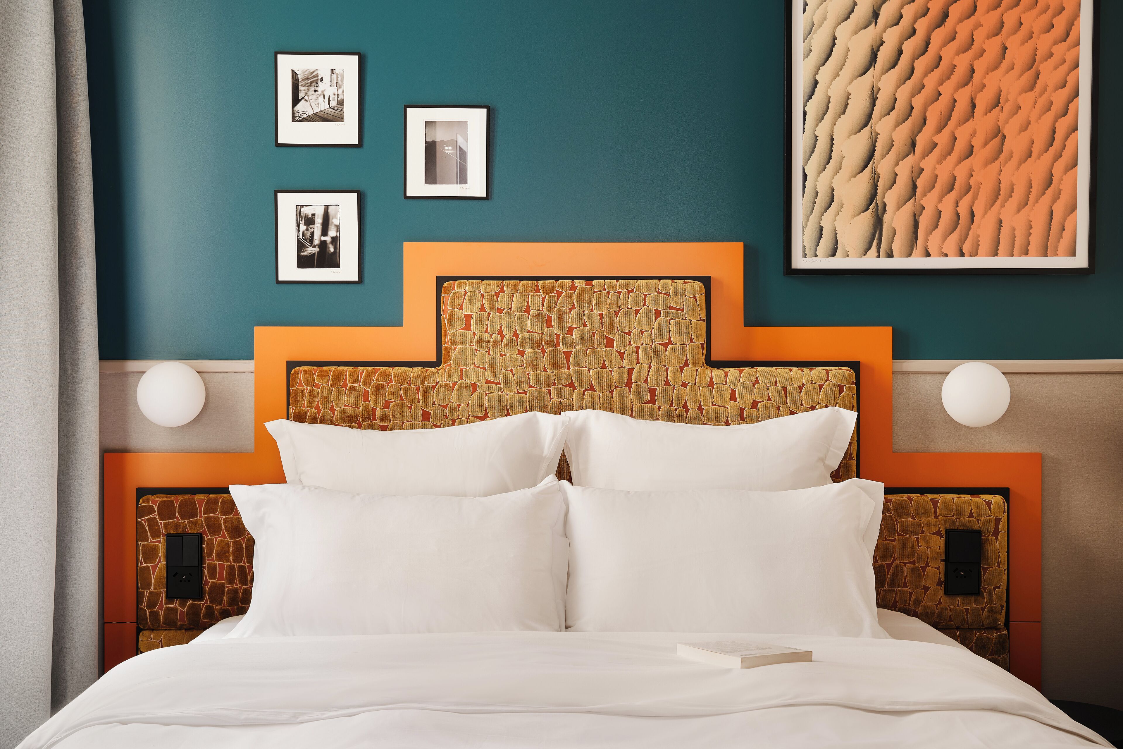 A neatly made bed with white linens, a headboard with a stone pattern, and a wall with framed pictures and a textured artwork.